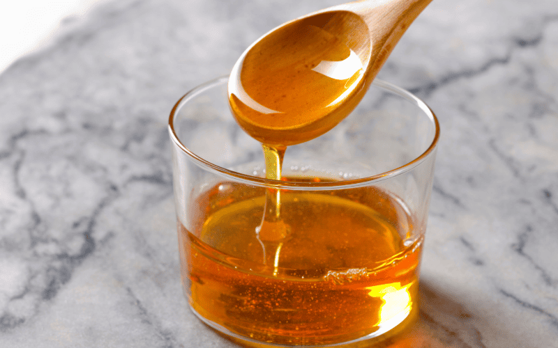 Best Substitute for Honey: 9 Alternatives to Sweeten Your Recipes Kelly Bejelly A transparent glass contains agave nectar, resting on a sleek marble counter, with a wooden spoon positioned above, releasing a gentle stream of agave nectar.