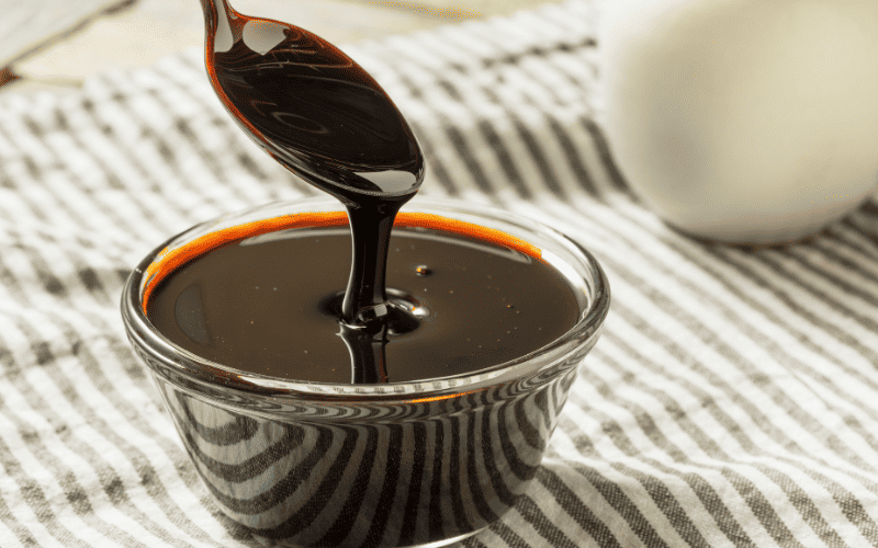 Best Substitute for Honey: 9 Alternatives to Sweeten Your Recipes Kelly Bejelly A glass brimming with rich molasses sits on a cloth, while a silver spoon hovers above, allowing thick molasses to elegantly drip from it; a milk jug's bottom is faintly visible in the background.