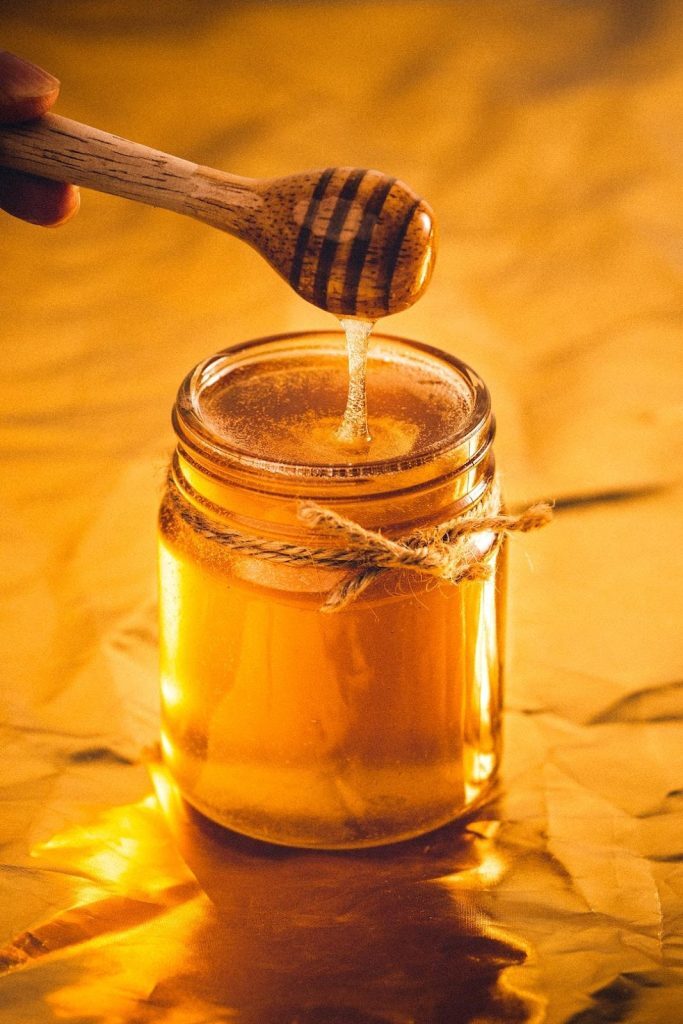 Best Substitute for Honey: 9 Alternatives to Sweeten Your Recipes Kelly Bejelly A glass jar of honey with a metal lid is placed on a flat surface. A wooden spoon is suspended a few inches above the jar, and a steady stream of golden honey flows from the spoon, creating a glistening arc as it descends into the jar. The backdrop features a soft, muted texture. The overall scene captures the process of honey being drizzled into the jar, showcasing its rich color and sticky fluidity.
