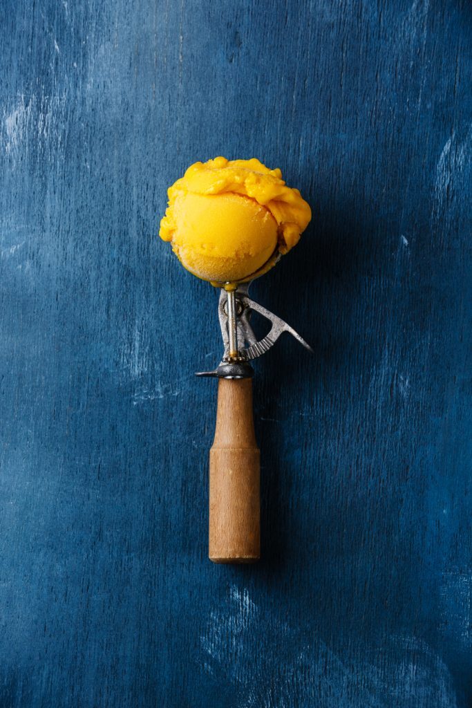 A ice cream scooper filled with a scoop of mango ice cream on a blue background 