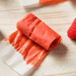 Homemade fruit roll ups: Easy Fruit leather recipe Kelly Bejelly Delicious fruit leather is artfully presented on parchment paper atop a wooden countertop, accompanied by a fresh, vibrant strawberry placed tastefully to the side.