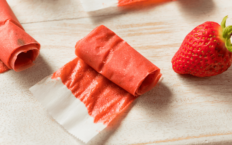 Recipe Index Kelly Bejelly Delicious fruit leather is artfully presented on parchment paper atop a wooden countertop, accompanied by a fresh, vibrant strawberry placed tastefully to the side.