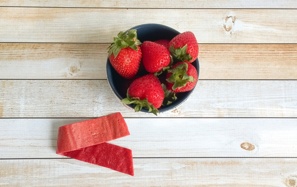 Homemade fruit roll ups: Easy Fruit leather recipe Kelly Bejelly Overhead view of a luscious strawberry fruit roll-up placed beside a bowl of fresh strawberries on a rustic wooden countertop.